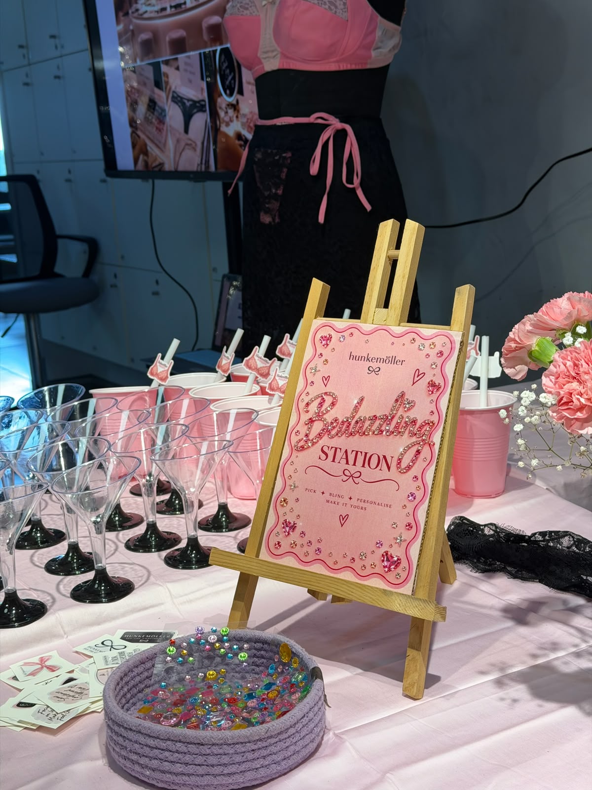 The Bedazzling Station easel sign on the activation table, surrounded by martini glasses, pink cups with bra-shaped stirrers, and a bowl of gems.